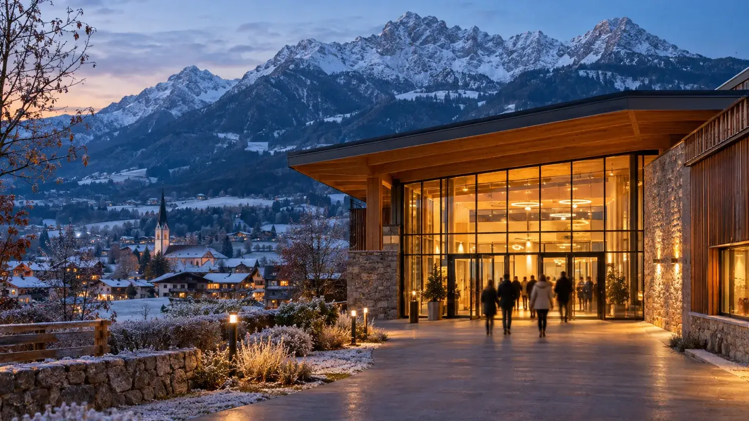 Beleuchteter Eingang einer Veranstaltungshalle an einem frühwinterlichen Abend, dahinter verschneite Berge