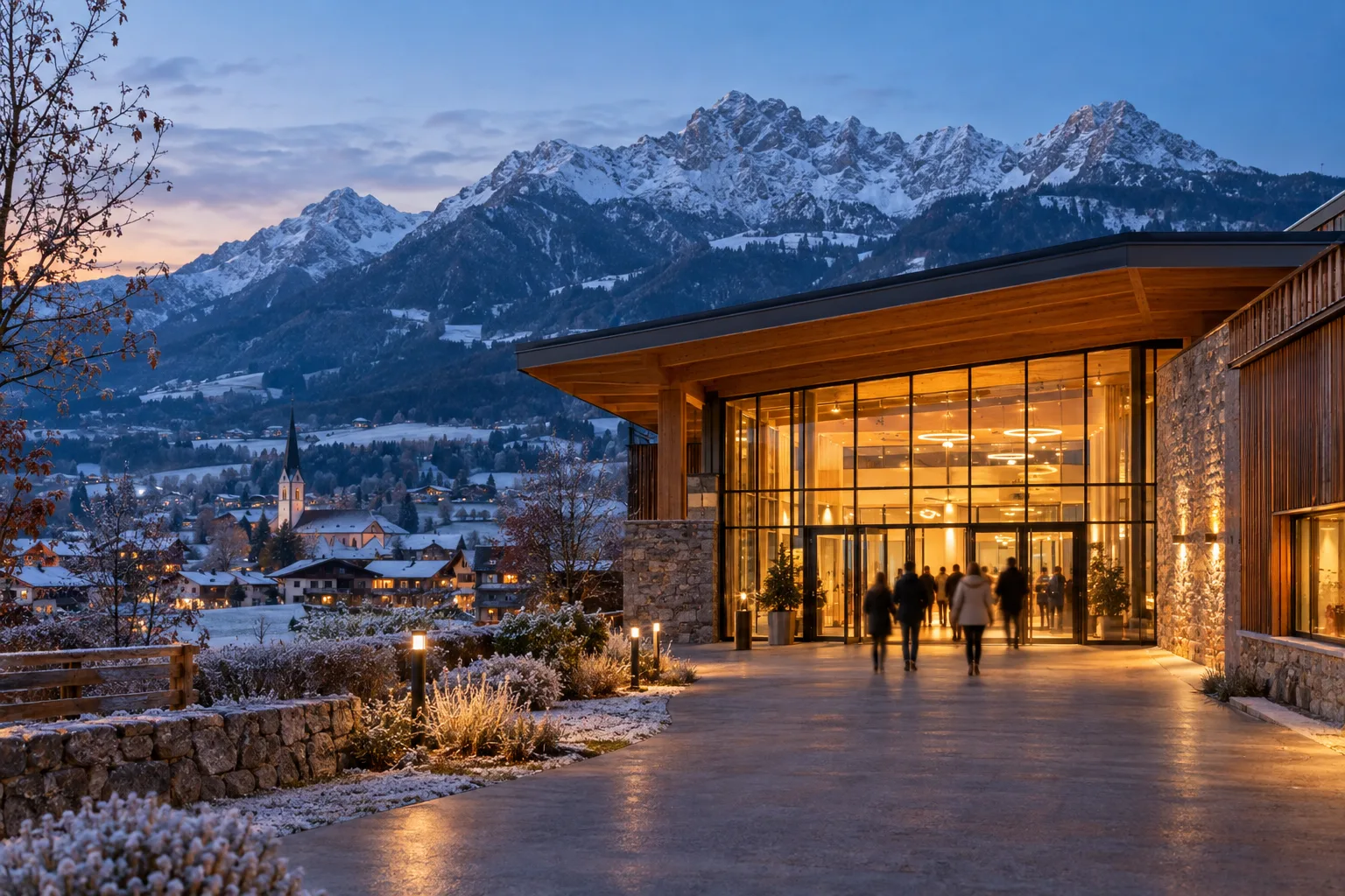 Beleuchteter Eingang einer Veranstaltungshalle an einem frühwinterlichen Abend, dahinter verschneite Berge